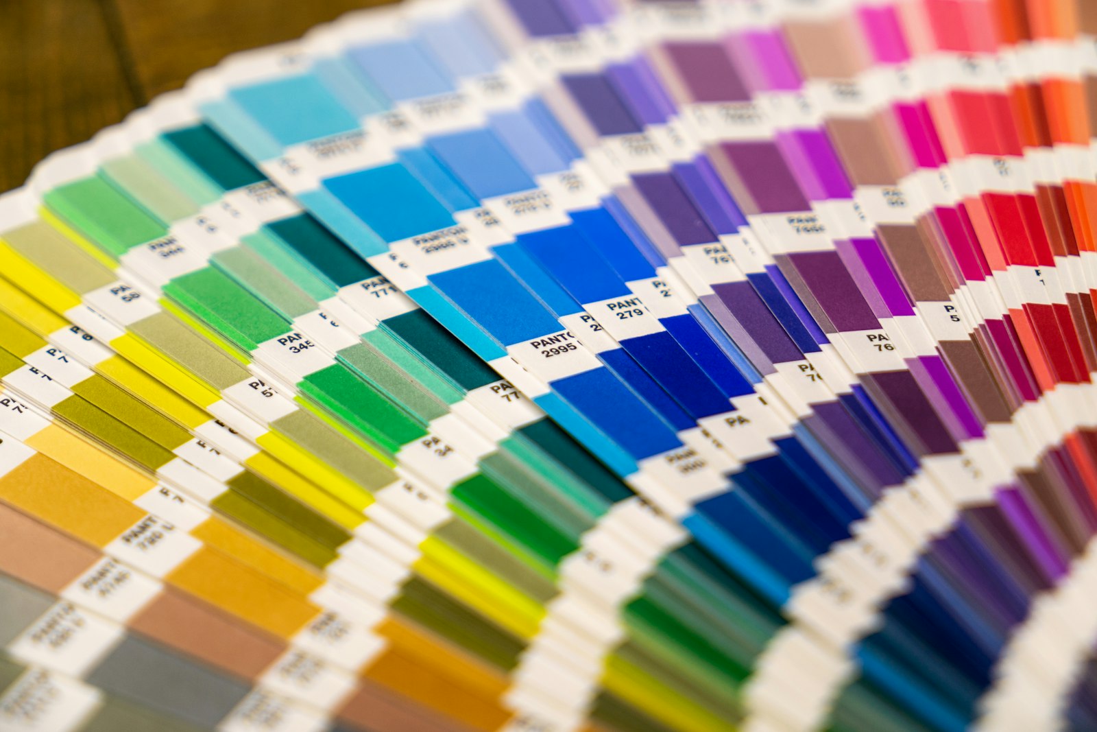A Pantone fan deck fanned open on a wooden table, hundreds of printed color chips running from yellow and green through blue to magenta and red