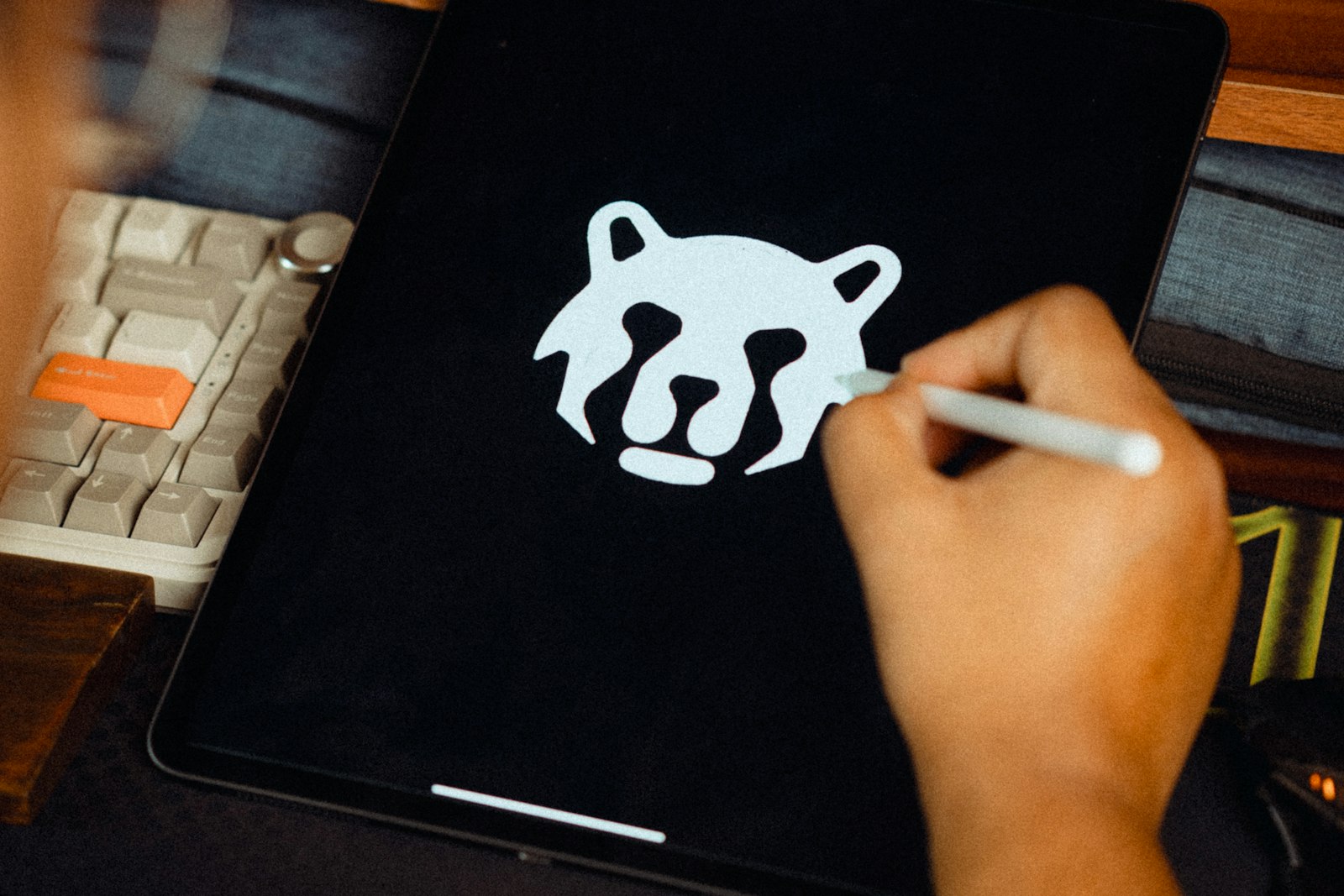 A hand with a stylus drawing a white bear head logo mark on a tablet screen, a mechanical keyboard on the desk beside it