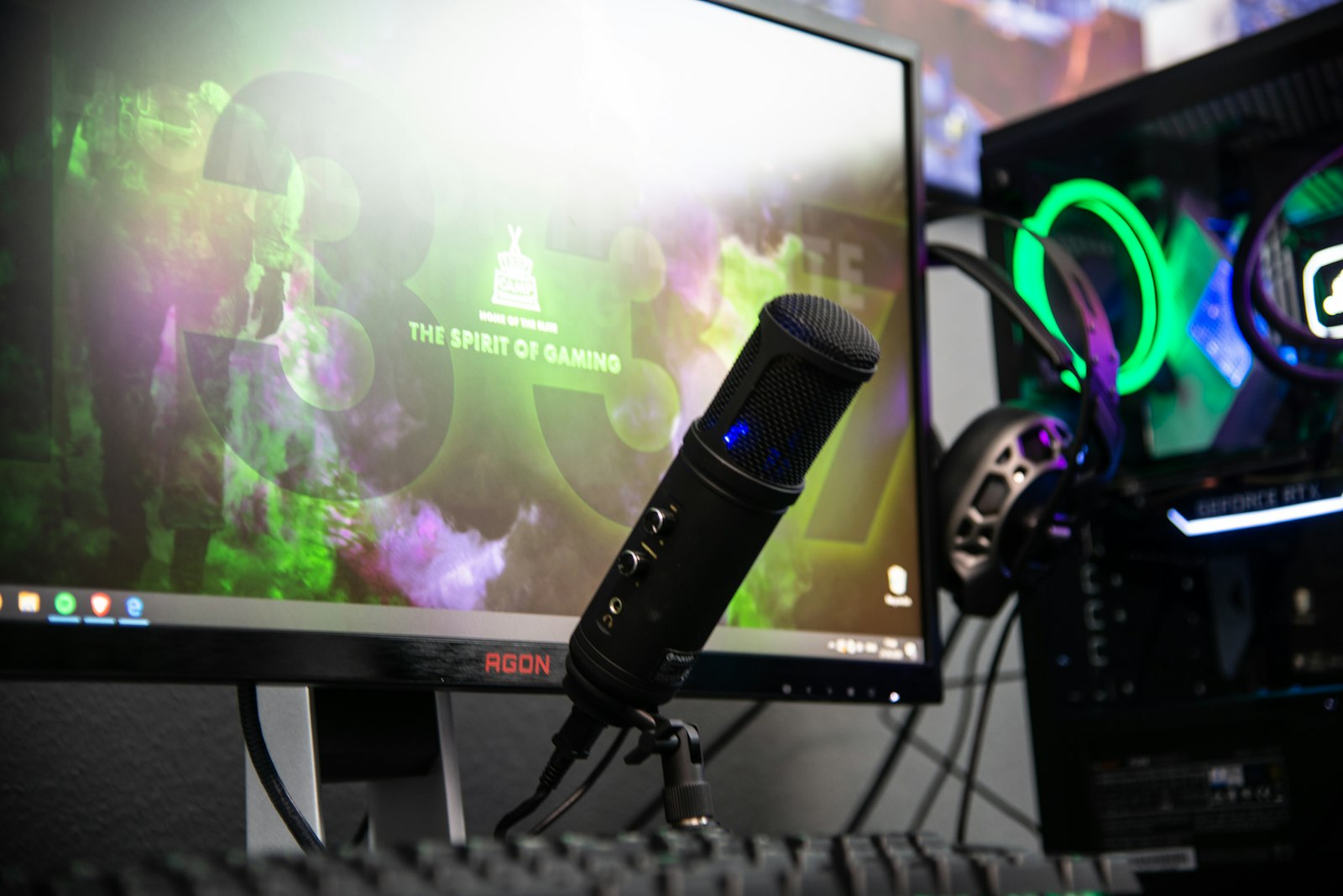 A streamer's desk with a USB condenser microphone in front of a monitor glowing with colorful graphics, an RGB-lit headset and gaming PC behind it, the kind of broadcast setup a music visualizer backdrop runs on