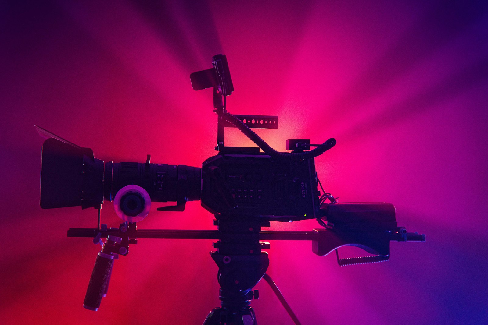 A rigged cinema camera on a tripod silhouetted against a backdrop lit deep pink and magenta, a music video set waiting for the artist